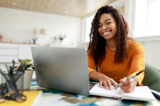 Woman sitting at desk, using computer and writing in notebook.