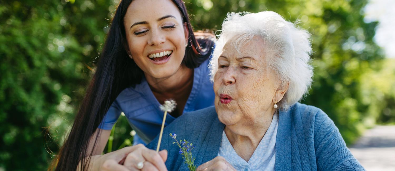 A younger caregiver in a blue uniform gently assists an older adult as they blow on a dandelion puff, sharing a joyful moment together in a lush green outdoor setting.