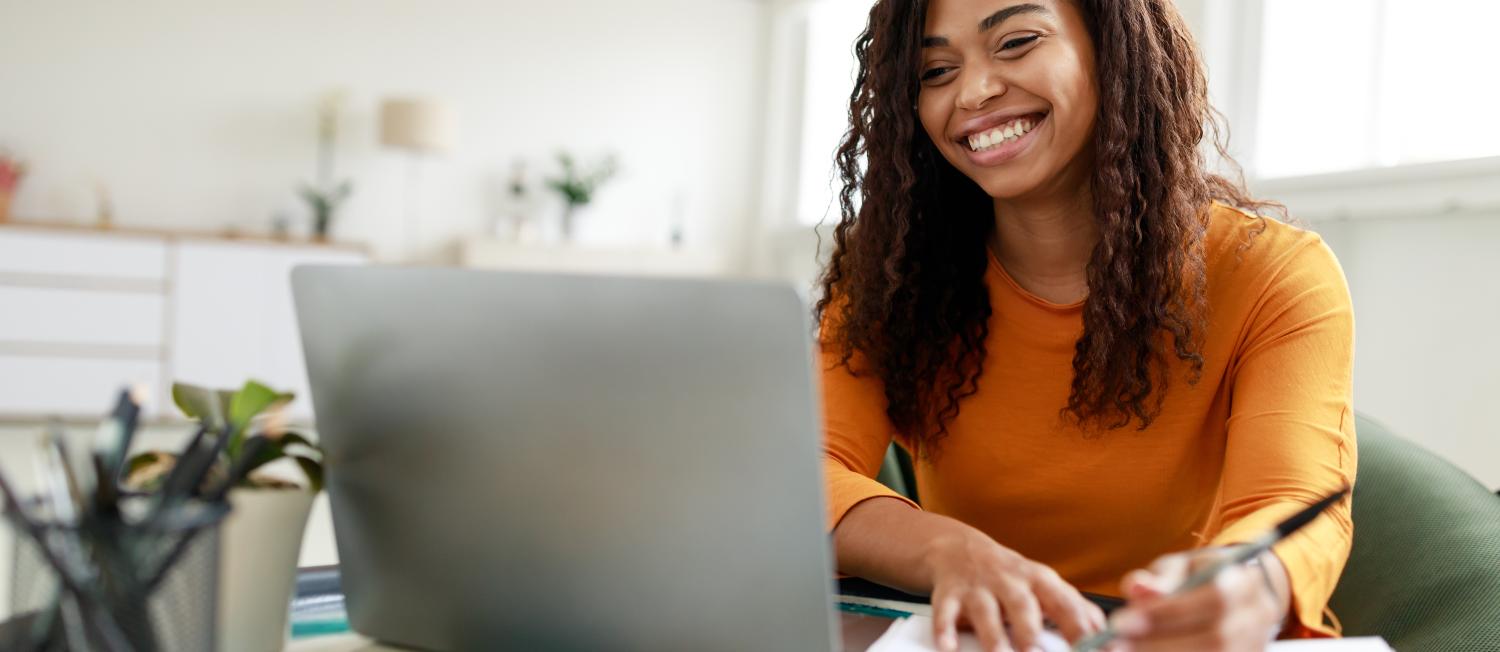 Woman sitting at desk, using computer and writing in notebook.