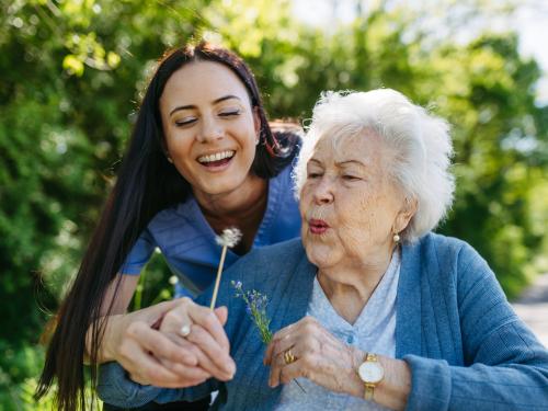 A younger caregiver in a blue uniform gently assists an older adult as they blow on a dandelion puff, sharing a joyful moment together in a lush green outdoor setting.
